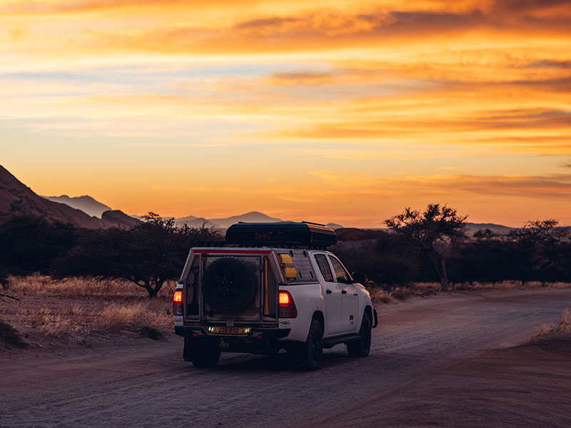 Asco Car Hire Autovermietung &ndash; Toyota Hilux 4x4-Mietwagen in Namibia mit Dachzelt, unterwegs auf einer namibischen Stra&szlig;e bei Sonnenuntergang